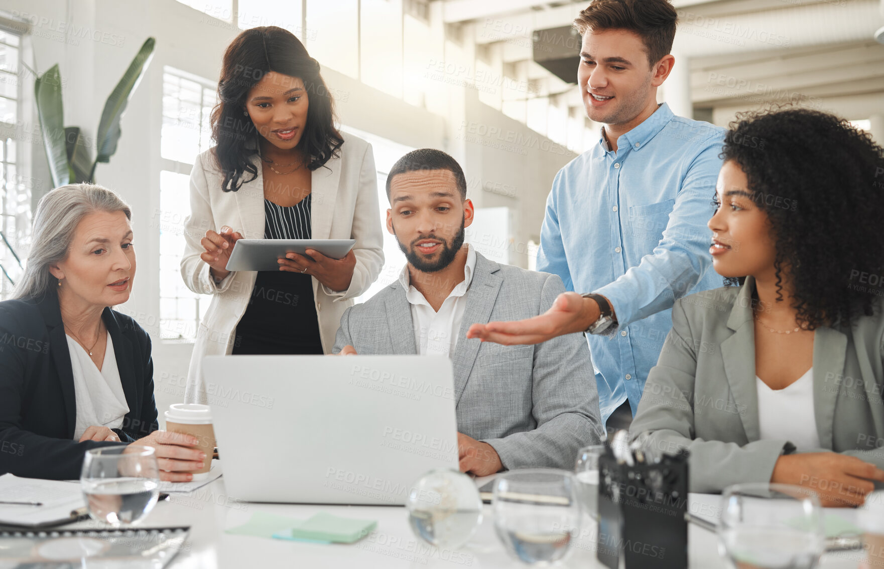 Buy stock photo Happy diverse group of businesspeople having a meeting while standing together at a table at work. Business professionals talking and planning while using a laptop in an office. Colleagues discussing a business strategy