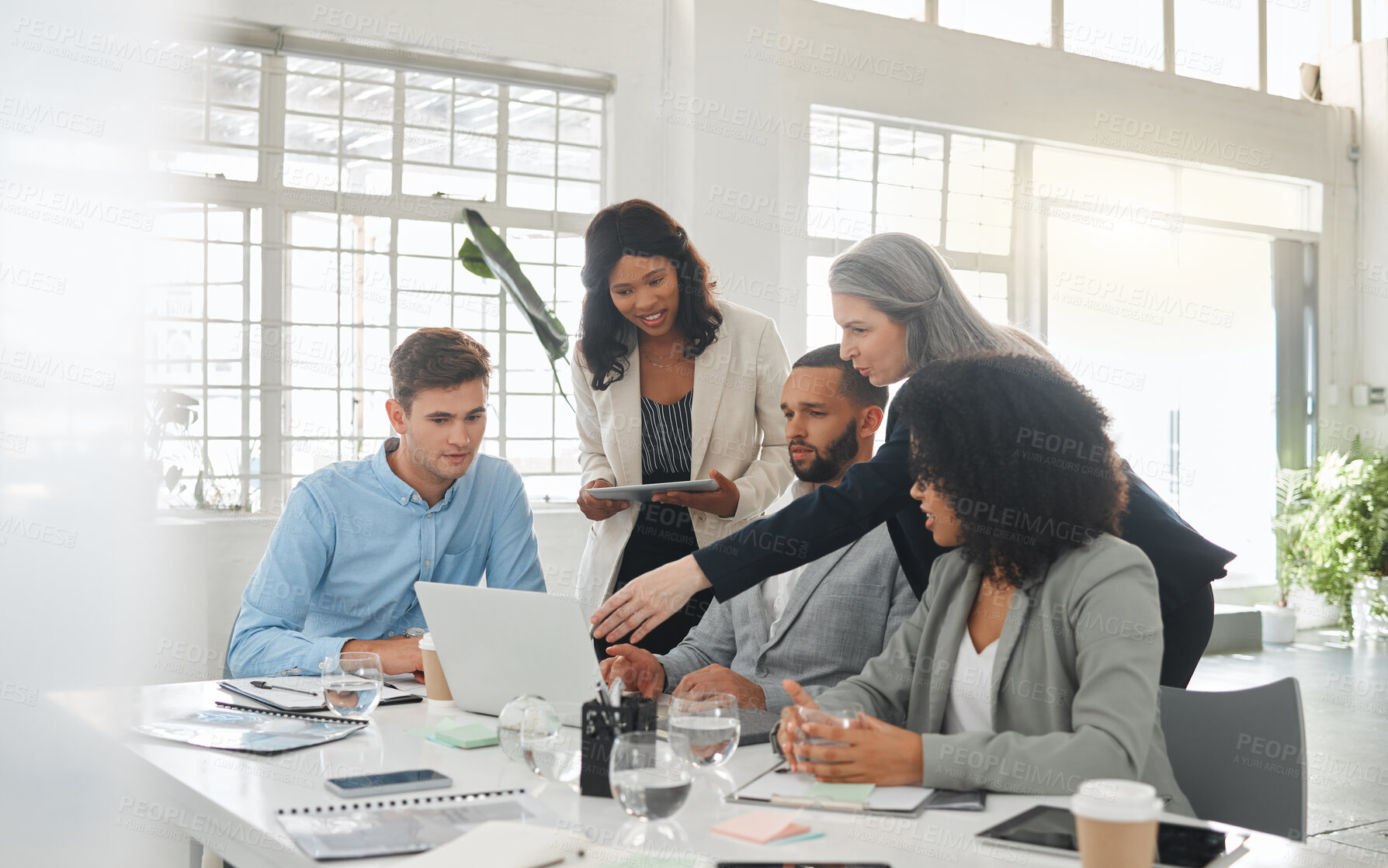 Buy stock photo Happy diverse group of businesspeople having a meeting while standing together at a table at work. Business professionals talking and planning while using a laptop in an office. Male and female colleagues discussing a business strategy