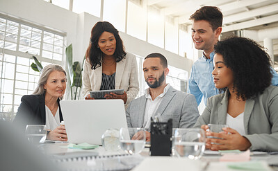 Happy diverse group of businesspeople having a meeting while standing together at a table at work. Business professionals talking and planning while using a laptop in an office. Male and female coworkers discussing a business strategy