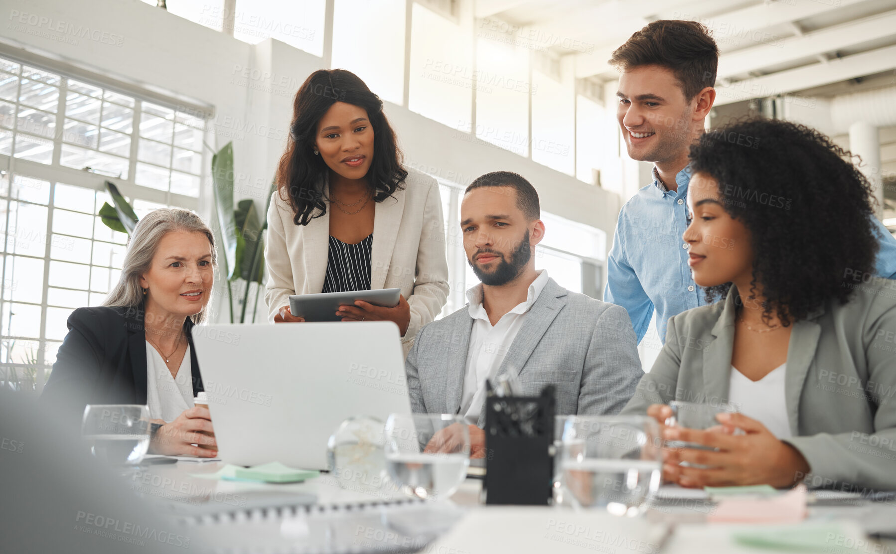 Buy stock photo Happy diverse group of businesspeople having a meeting while standing together at a table at work. Business professionals talking and planning while using a laptop in an office. Male and female coworkers discussing a business strategy