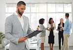 Young focused mixed race businessman reading a form on clipboard while standing in an office with colleagues. Serious confident hispanic male boss checking a document at work
