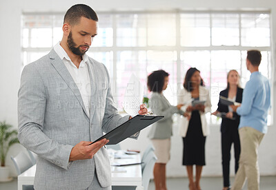 Buy stock photo Young focused mixed race businessman reading a form on clipboard while standing in an office with colleagues. Serious confident hispanic male boss checking a document at work