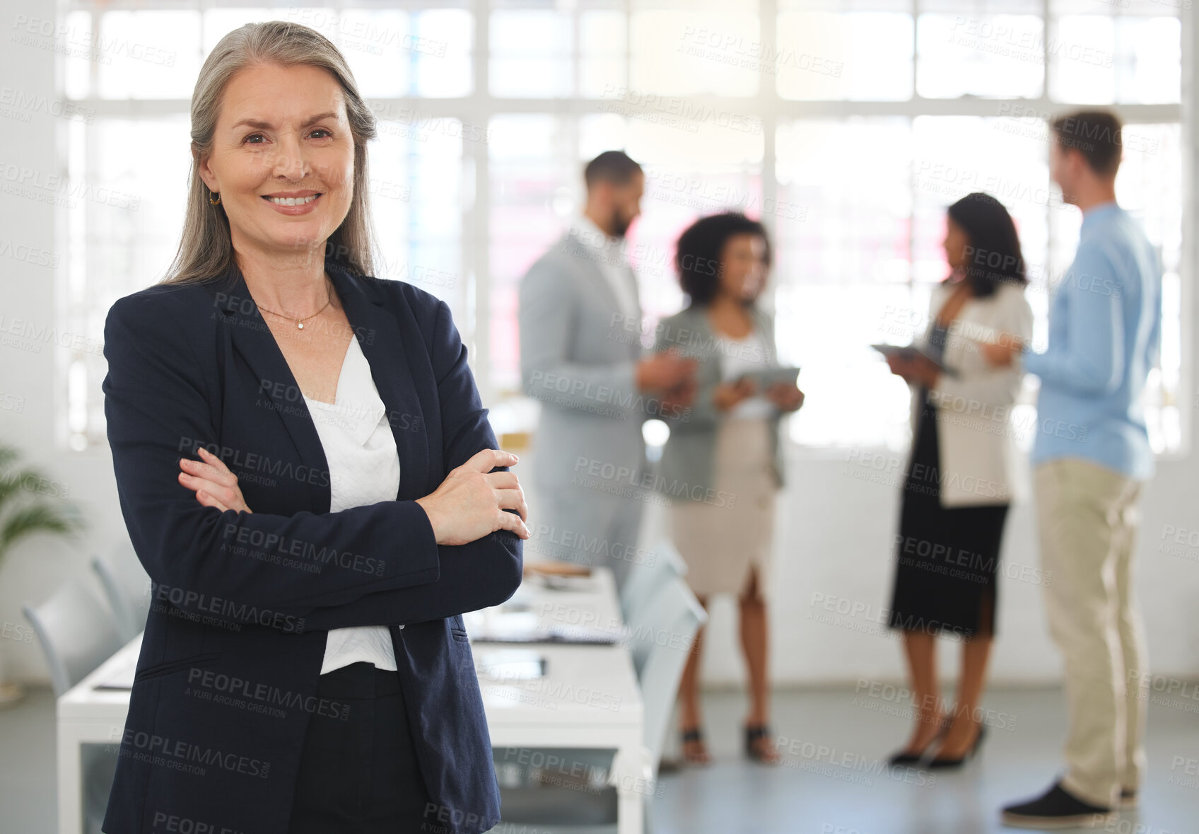 Buy stock photo Mature caucasian businesswoman standing with her arms crossed while in an office with colleagues. Happy female boss In a meeting with coworkers