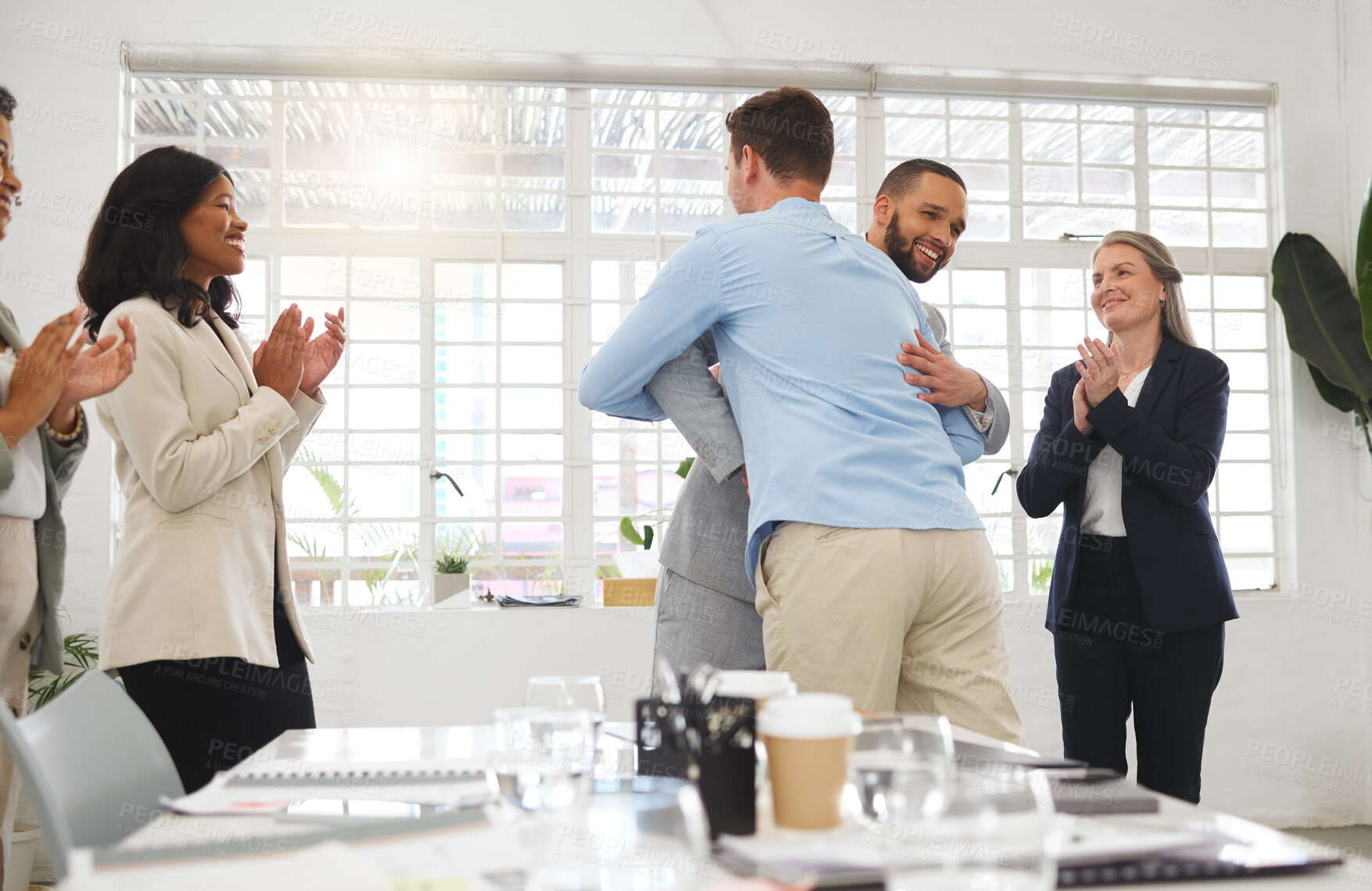 Buy stock photo Businessmen hugging each other in support and unity while colleagues clap hands in a meeting at work. Diverse group of cheerful businesspeople welcoming an employee to their workplace together