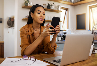 Young mixed race woman drinking a cup of coffee while using her phone and working on a laptop alone in the morning in the kitchen. Content hispanic female smiling and enjoying a cup of tea while reading an email on her cellphone at home