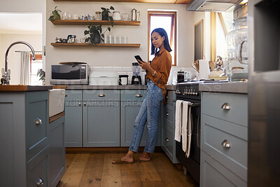 Young mixed race woman drinking a cup of coffee while using her phone alone in the morning in the kitchen. One hispanic female enjoying a cup of tea while using social media on her phone at home