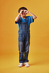 Curious little boy looking through fingers shaped like binoculars while wearing casual clothes against a yellow studio background