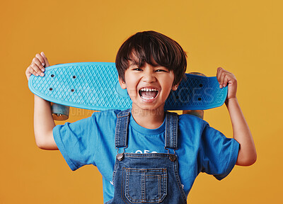 An adorable little asian boy looking happy while holding his skateboard against an orange background. Cute boy wearing casual clothes cheering as he carries his skateboard. Cute child having fun