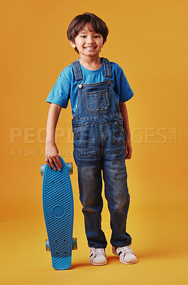 An adorable little asian boy looking happy while holding his skateboard against an orange background. Cute boy wearing casual clothes smiling as he holds his skateboard. Cute child having fun