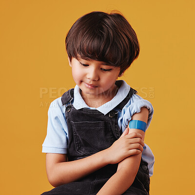 A cute little asian boy holding his arm and showing his bandaid. Child wearing a plaster after being forced to get the vaccine, sad kid after getting an injection against an orange background