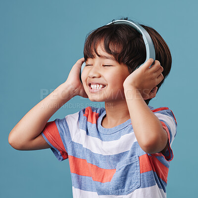 A cute young asian boy enjoying listening to music from his headphones. Adorable Chinese kid feeling the magic of music while posing against a blue studio background