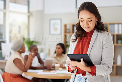 Smiling asian business woman using a digital tablet while colleagues sit behind her in office. Ambitious and happy professional standing and browsing schedule on technology before leading a meeting
