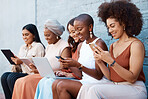 Group of five young happy cheerful businesswomen sitting on a bench against a wall outside in the city and using tech. Businesspeople smiling using technology and sitting in a row outdoors