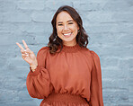 One young business woman of mixed race ethnicity standing outside against a grey wall and gesturing the peace sign with her hand. Happy and confident female executive looking positive and successful
