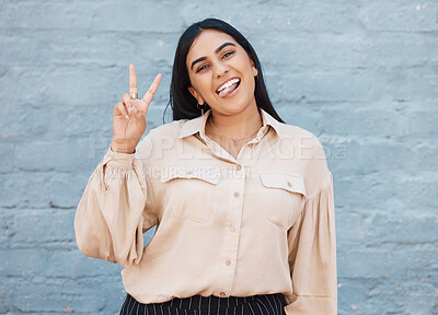 One young business woman of indian ethnicity standing outside against a grey wall and gesturing the peace sign with her hand. Happy and confident mixed race executive looking positive and successful