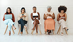 Group of five young businesswomen sitting against a wall in an office and using tech. Businesspeople using technology and sitting in a row together inside at work