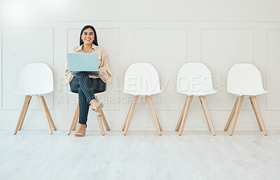 Young happy smiling mixed race hispanic businesswoman working on laptop while waiting for interview sitting on a chair against a white wall in an office at work
