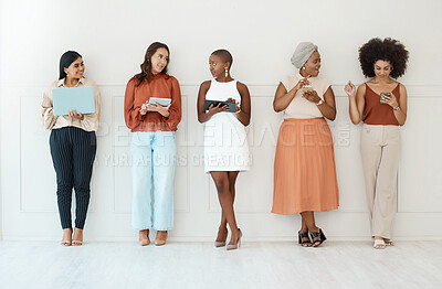 Group of five young happy cheerful businesswomen standing against a wall in an office and using tech. Happy colleagues talking and using technology while standing in a row together at work