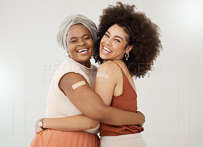 Two young friends wearing and showing a bandaid on their arms standing against a white wall together at work. Happy businesswomen hugging with a plaster on their arms after getting the covid vaccine
