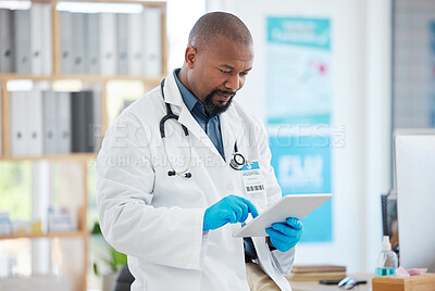 Focused doctor using a digital tablet in his clinic office. African american specialist using an online device. Caring gp wearing gloves using a wireless digital device.