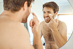 One topless handsome man applying cream or moisturiser to his face in a bathroom at home. Caucasian male using a lotion or sunscreen while looking in a mirror in his apartment