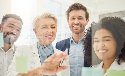 Mature caucasian businesswoman mentoring her diverse team while planning a project and analysing notes on a glass wall in an office. Group of smiling businesspeople brainstorming ideas and strategies together