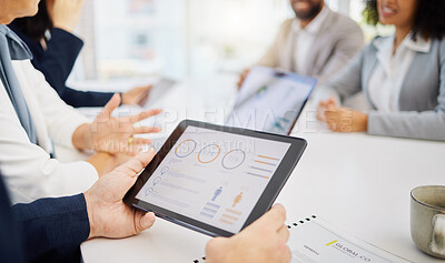 Closeup of a businessman analysing research stats and marketing data on a digital tablet device in an office with his colleagues in the background. Entrepreneur reviewing infographics during a meeting with his team