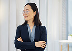 One confident young asian businesswoman wearing glasses and looking thoughtful while standing with her arms crossed in an office. Happy ambitious entrepreneur daydreaming about success in her startup
