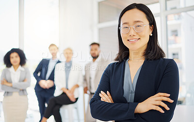 Portrait of a confident young asian businesswoman wearing glasses and standing with her arms crossed in an office with her colleagues in the background. Ambitious entrepreneur and determined leader ready for success in her startup with her team