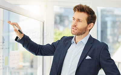 One focused young caucasian businessman writing ideas on a glass wall while planning a project in an office. Ambitious entrepreneur brainstorming a solution and marketing strategy while presenting a workshop to his team