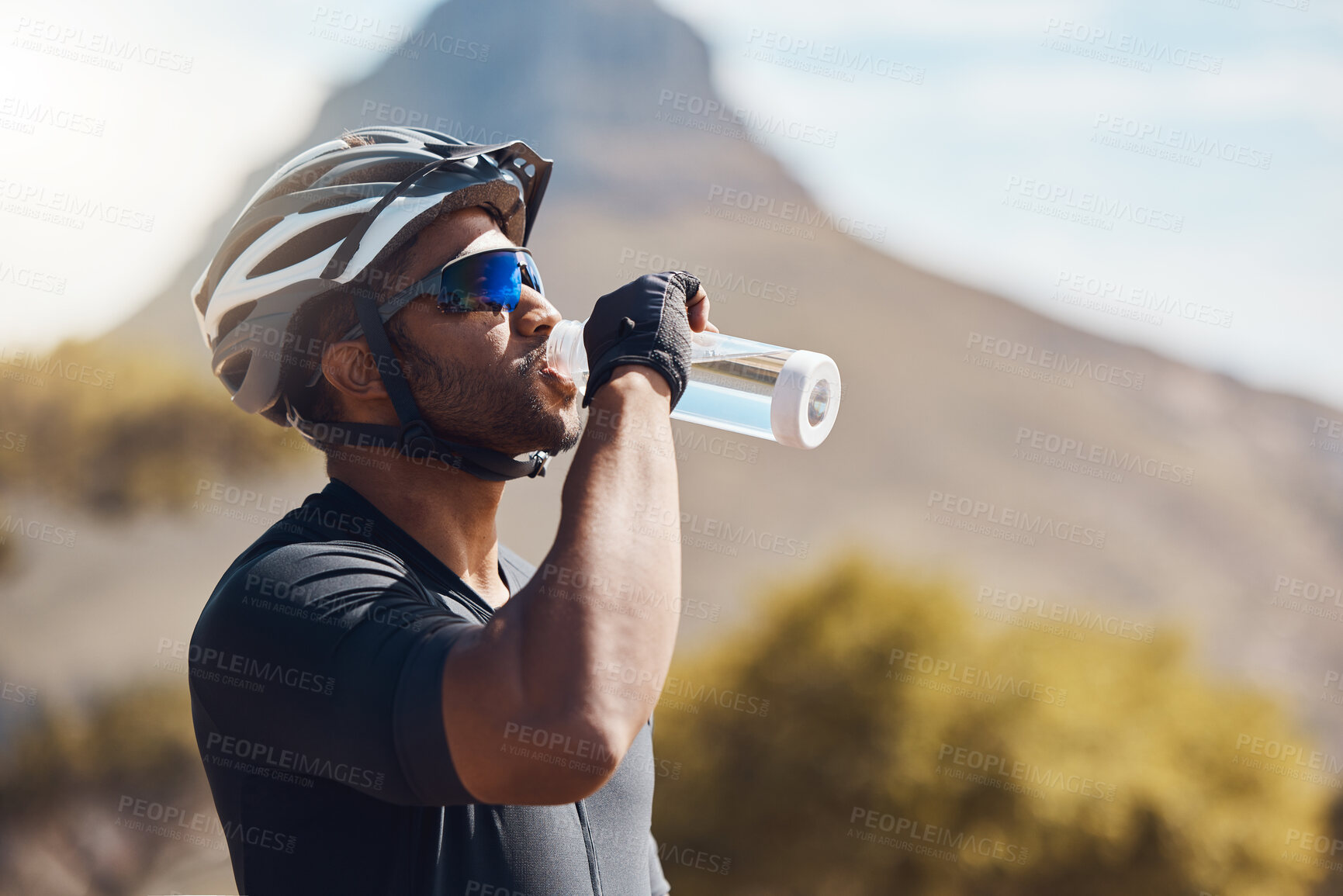 Buy stock photo Close up of male cyclist taking a break and drinking water from a bottle. Fit young man wearing glasses and a helmet while drinking water and standing outside. Athletic man training in nature environment 