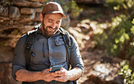 Happy caucasian man using smartphone while carrying a backpack and hiking alone in the mountains during the day. Smiling fit and active man using mobile app or messaging while exercising