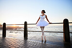 Elegant young ballerina wearing white fancy dress and pumps while leaning against a railing on the promenade with a beautiful ocean background at sunset 