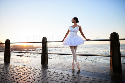 Elegant young ballerina wearing white fancy dress and pumps while leaning against a railing on the promenade with a beautiful ocean background at sunset