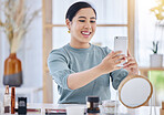 Cheerful young woman sitting at her desk taking a selfie on her cellphone before applying skincare products. Smiling woman using her smartphone to take photos while doing her beauty treatment routine