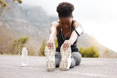 One african american female athlete with an afro listening to music on her earphones while exercising outdoors in nature. Dedicated black woman smiling while warming up before a workout outside