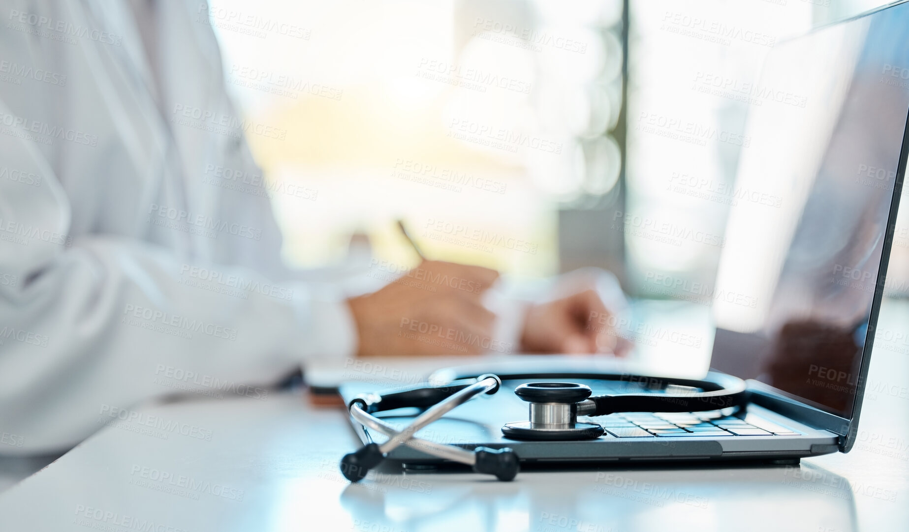 Buy stock photo Closeup of a stethoscope lying on a laptop on a desk while a doctor writes a prescription in the background. Medical professional writing notes, planning with a laptop and stethoscope in the office
