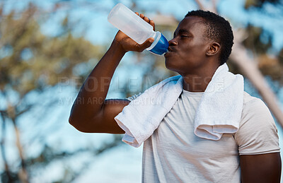 Young active black male athlete holding a bottle and drinking water with a towel around his neck in a forest outside. Runny feeling dehydrated quenching his thirst with some water on sunny hot summers day