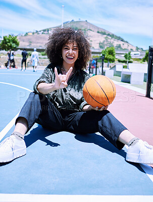 Cheerful young hispanic woman making the rock gesture with her hands and fingers, holding a basketball on the court. Happy African American woman with afro playing basketball with crazy expression