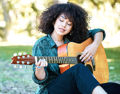 Young mixed race woman with a curly afro sitting in a park and playing the guitar alone. Young happy hispanic woman playing the guitar while sitting in nature relaxing and playing the guitar
