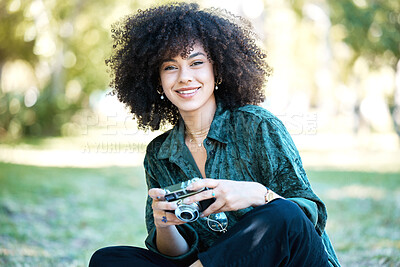 Smiling young photographer enjoying a day in the park sitting on the grass taking photos using her digital camera. Happy hispanic woman with a curly afro holding her camera equipment in the garden