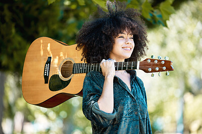 Happy smiling young mixed race female musician holding her guitar while standing snd enjoying a relaxing day in the park alone. Hispanic woman with a curly afro playing her guitar in nature alone