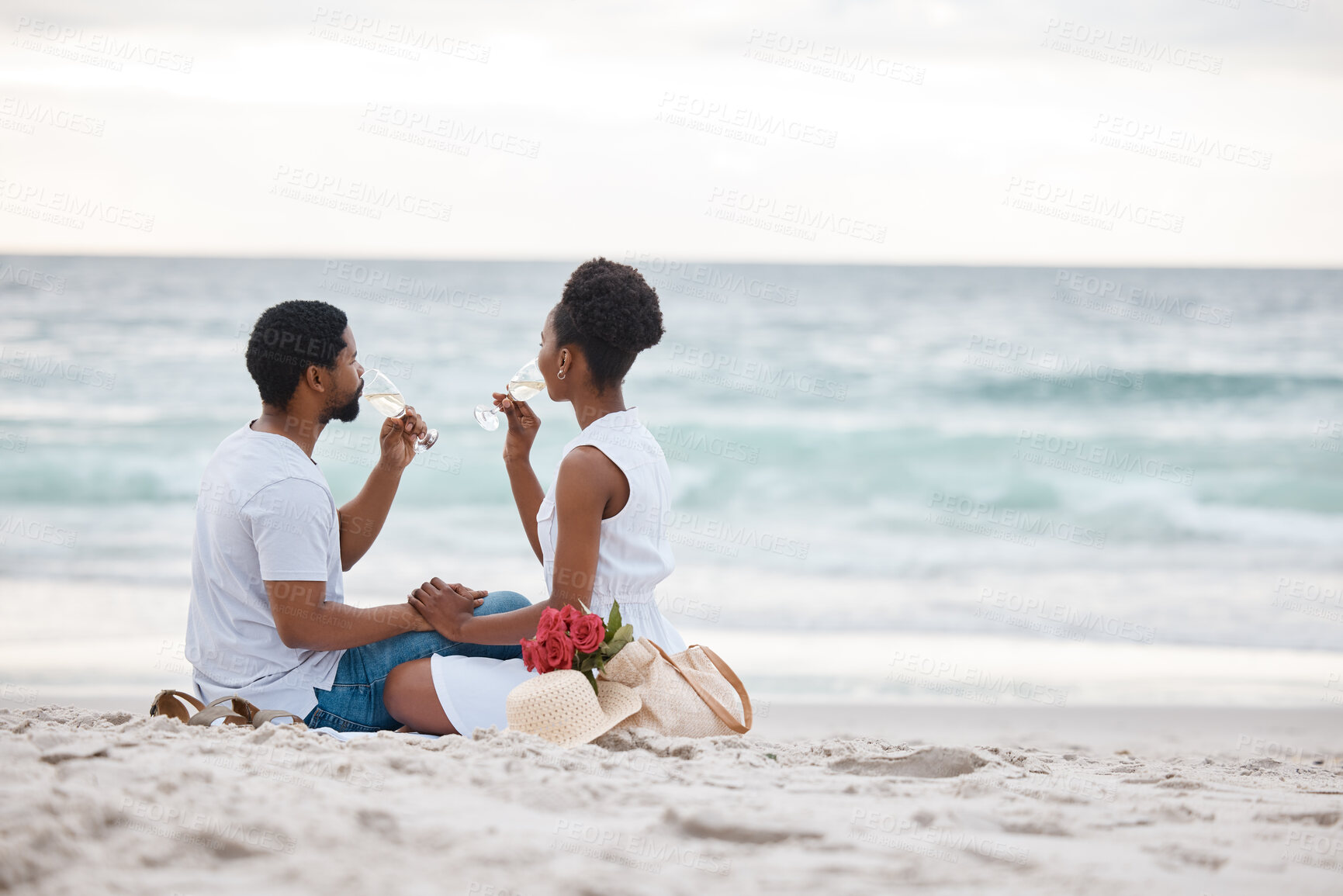Buy stock photo Young african american couple drinking champagne at the sea together. Caring boyfriend and girlfriend spending a day on the beach. Loving husband and wife having a drink on the beach