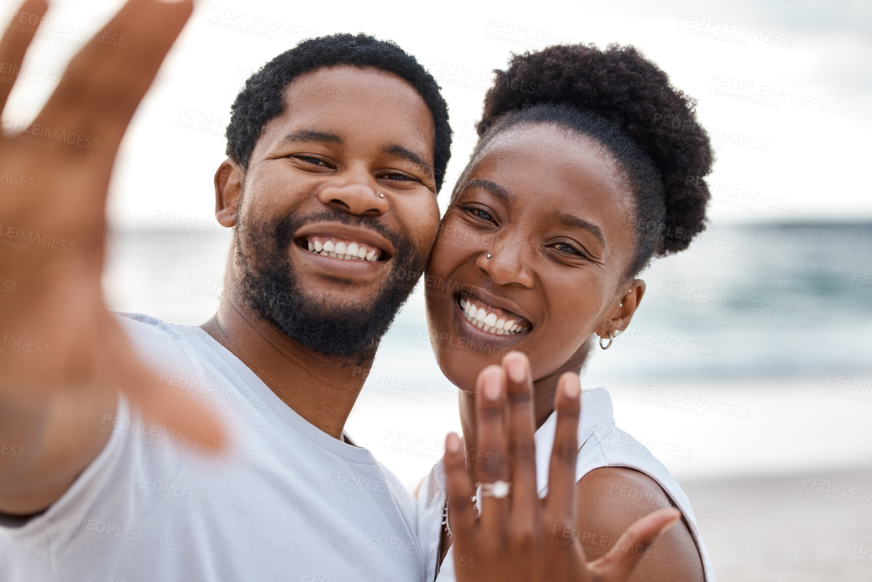 Buy stock photo Portrait of an african american couple taking a selfie while showing a ring at the sea together. Happy boyfriend and girlfriend taking a photo after eating engaged on the beach