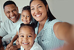 Photo of a loving mother holding phone and taking selfie or recording funny video with her family at home. Happy mixed race family with two children and parents posing together for a family picture on a mobile phone