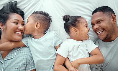 Happy funny mixed race family with two children relaxing and lying together on a bed at home, from above. Adorable little boy kissing his mother on the cheek while his sister and father look at each other and laugh
