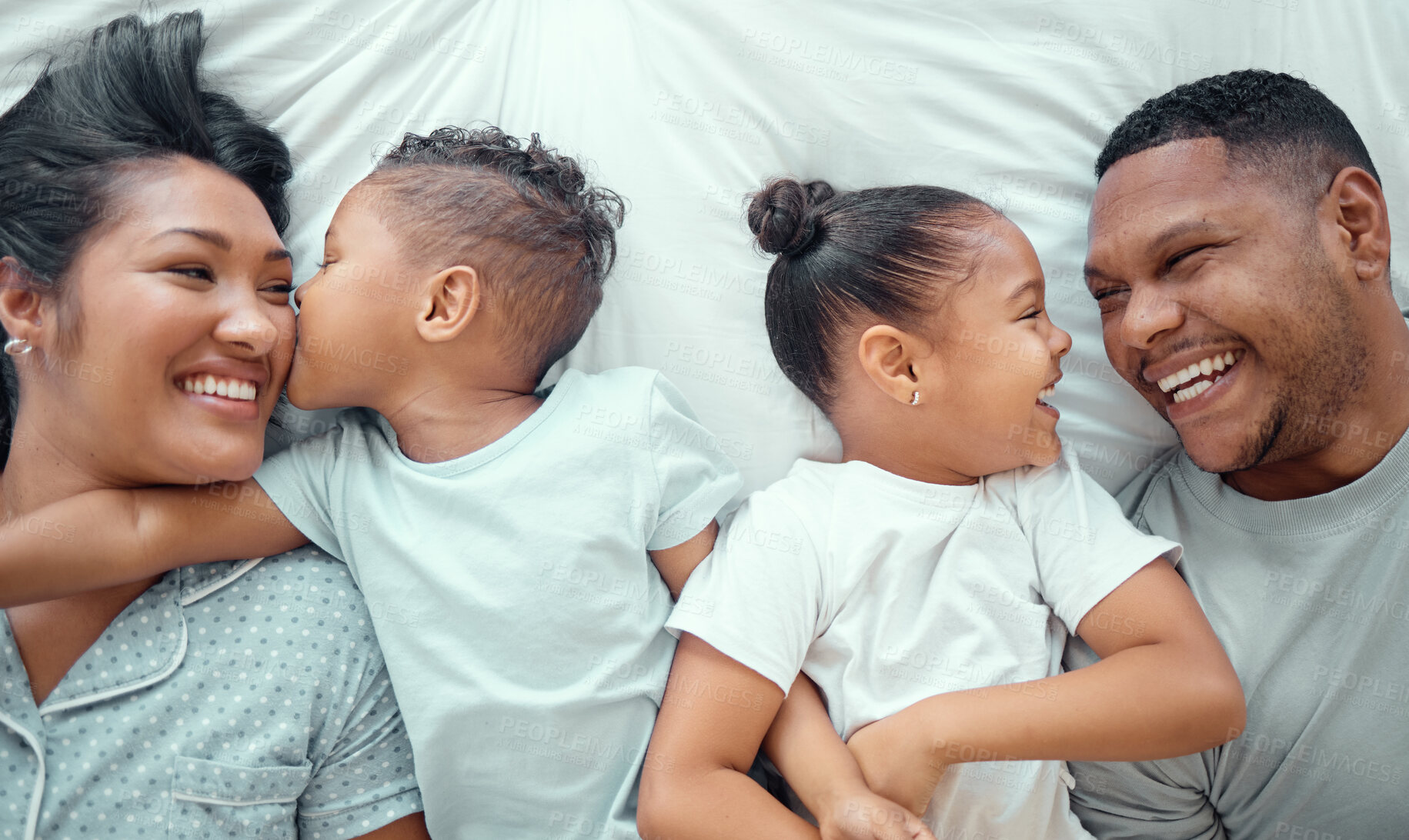 Buy stock photo Happy funny mixed race family with two children relaxing and lying together on a bed at home, from above. Adorable little boy kissing his mother on the cheek while his sister and father look at each other and laugh