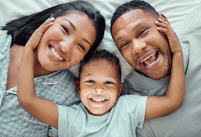 Buy stock photo Portrait of adorable little boy with his hands on his parents paces pulling them close while lying on a bed. Cute son lying in between his mother and father, from above. Faces of loving parents bonding with their son