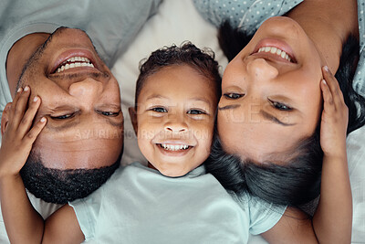 Close up portrait of adorable little boy with his hands on his parents paces pulling them close while lying on a bed. Cute son lying in between his mother and father, from above. Faces of loving parents bonding with their son