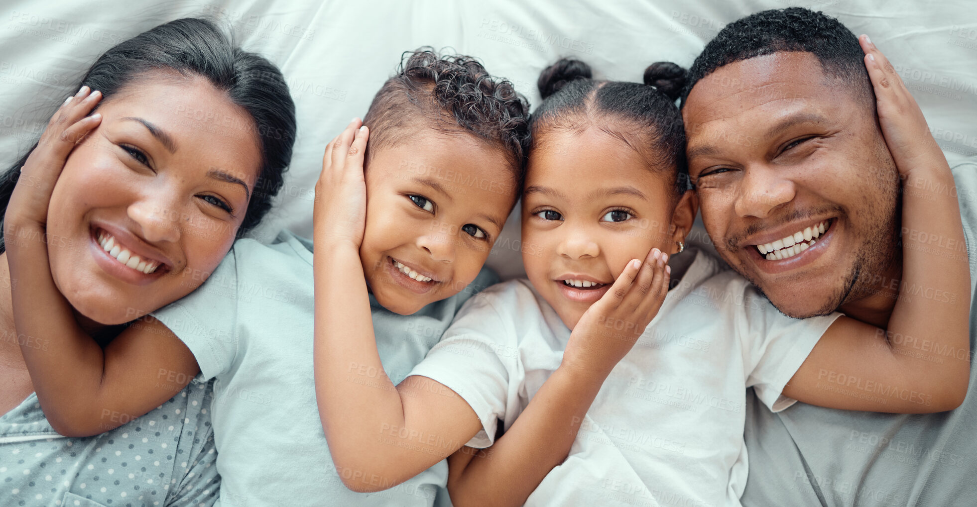 Buy stock photo Portrait of happy mixed race family with two children relaxing and lying together on a bed at home, from above. Little brother and sister with their hands on each others faces while bonding with their loving parents